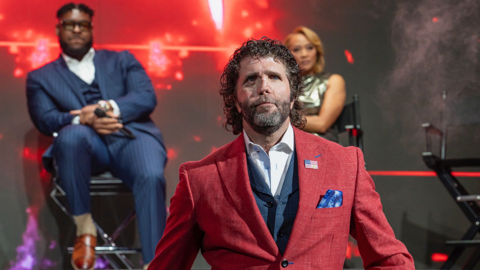 A man in a red suit with an American flag pin stands on stage, flanked by a man in a blue suit and a woman, against a vibrant red background.