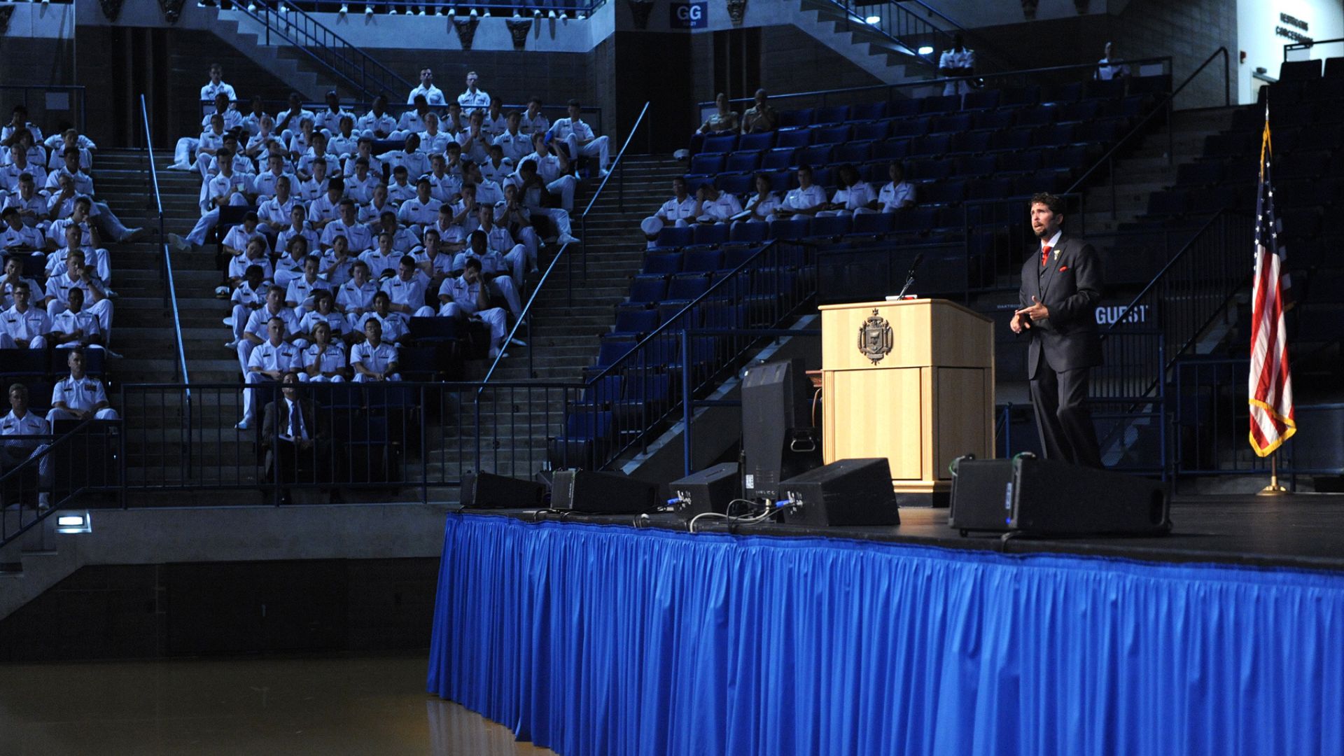 Image of a man speaking at a podium on a stage with a large audience in uniform, with an American flag in the background, highlighting a public speaking event.