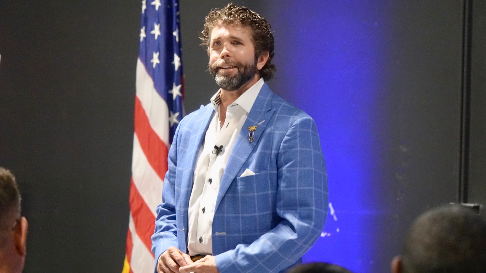 A man in a blue suit with medals speaking on stage, standing in front of an American flag with an audience in the background.