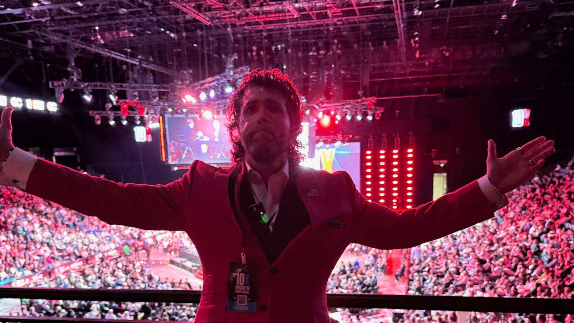 A man in a red suit with arms outstretched on stage at a vibrant 10X event, addressing a large audience under colorful lights.