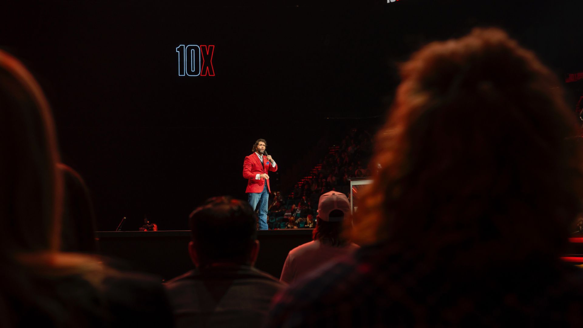 Image of a man in a red jacket speaking on stage with a "10X" sign, addressing an audience, emphasizing public speaking and communication skills.