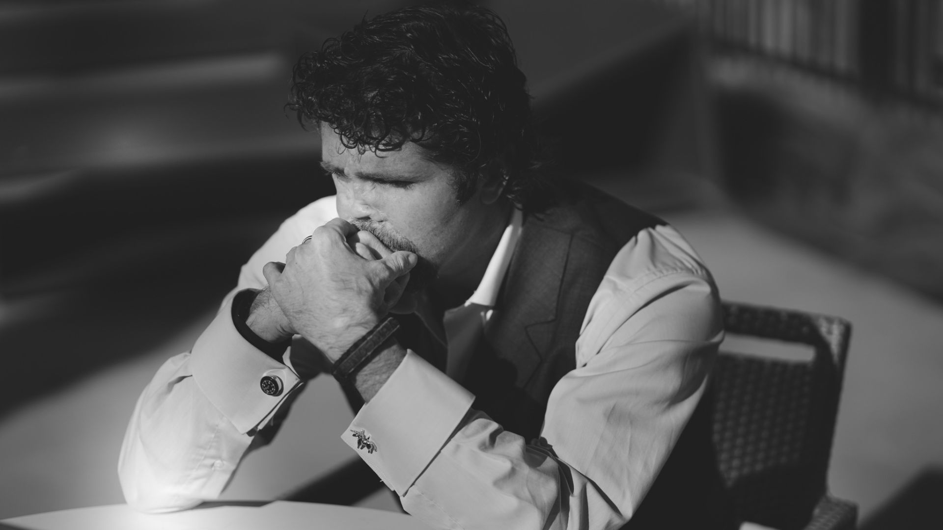 Black-and-white photo of a man in a suit and vest, sitting with hands near face, showcasing cufflinks and a contemplative pose.