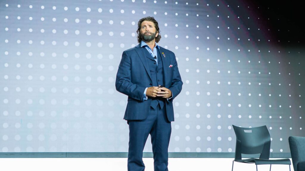 Image of a man in a blue suit and red sneakers standing on a stage with a dotted backdrop, likely speaking at an event.