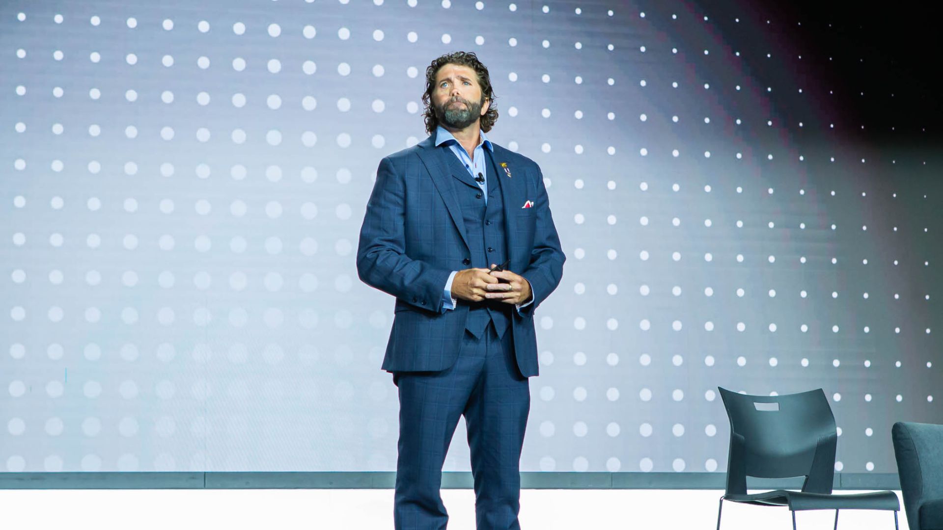 Image of a man in a blue suit and red sneakers standing on a stage with a dotted backdrop, likely speaking at an event.