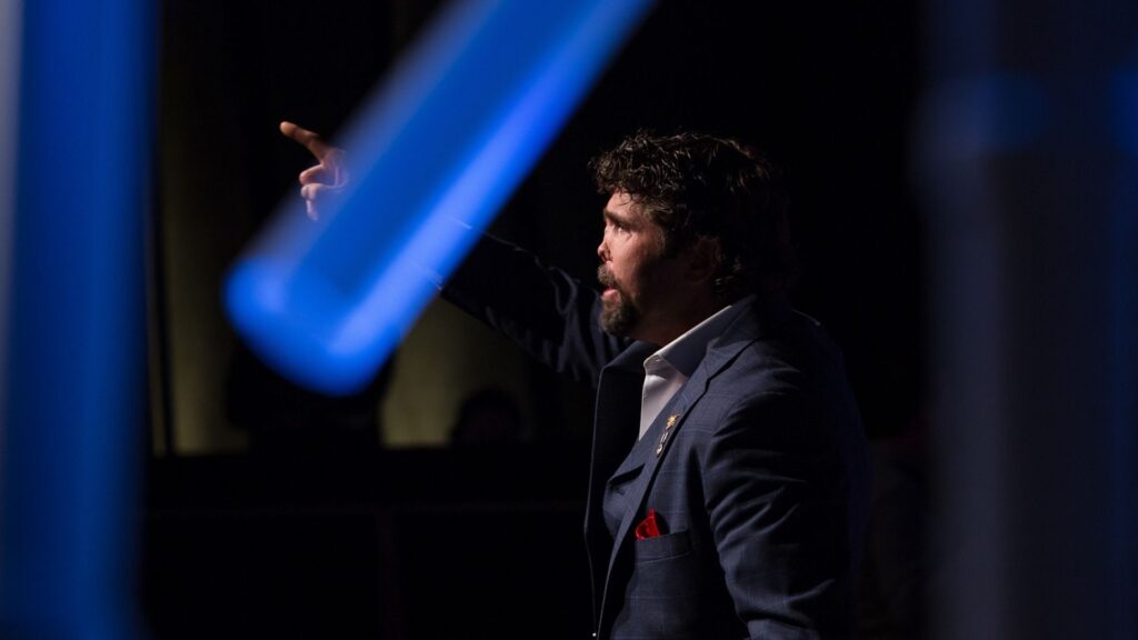 Image of a man in a suit with a red pocket square, pointing upward on a stage with blue lighting, emphasizing a motivational speaking moment.