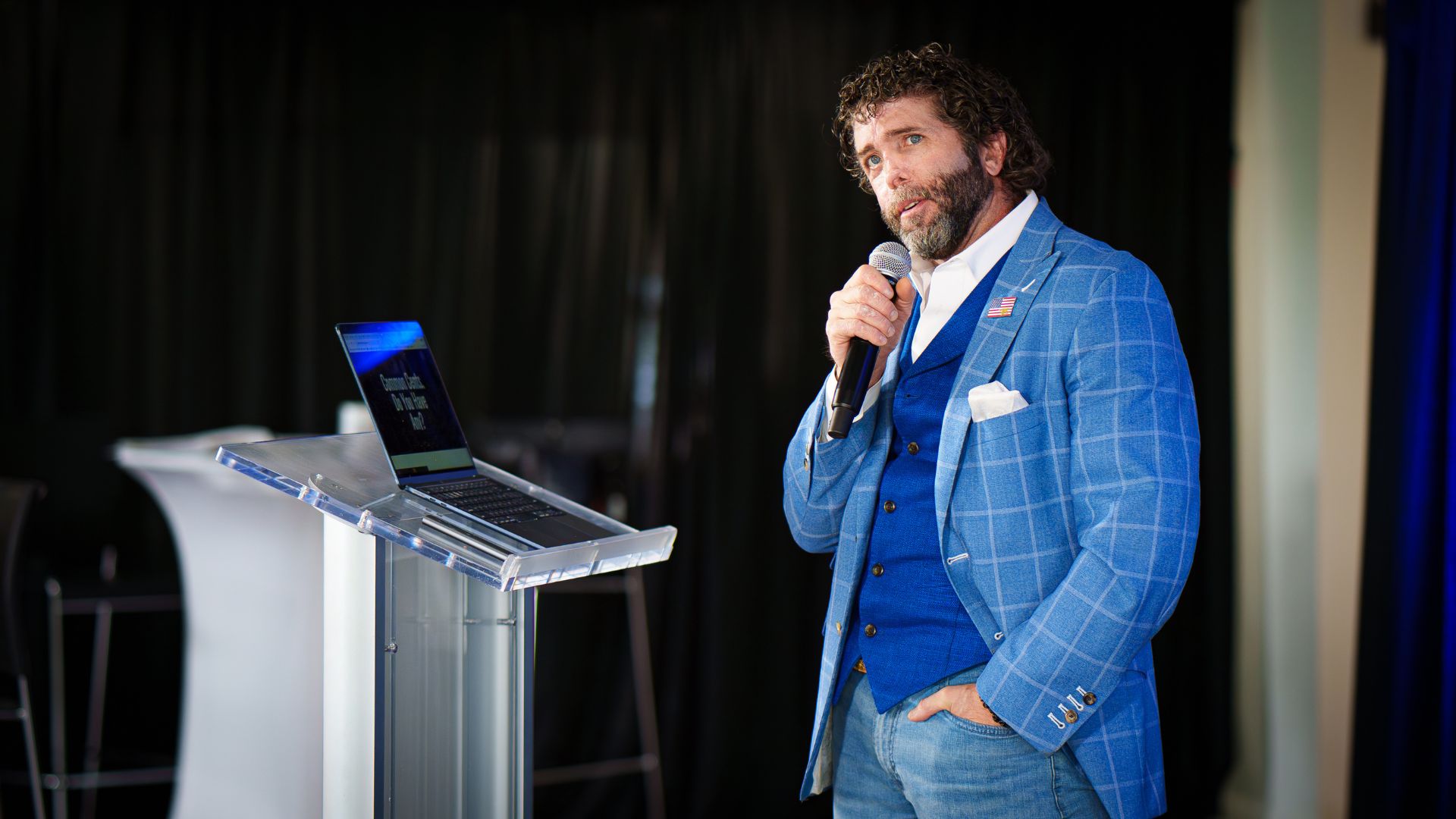 A speaker in a blue suit holding a microphone at a podium with a laptop, presenting confidently.