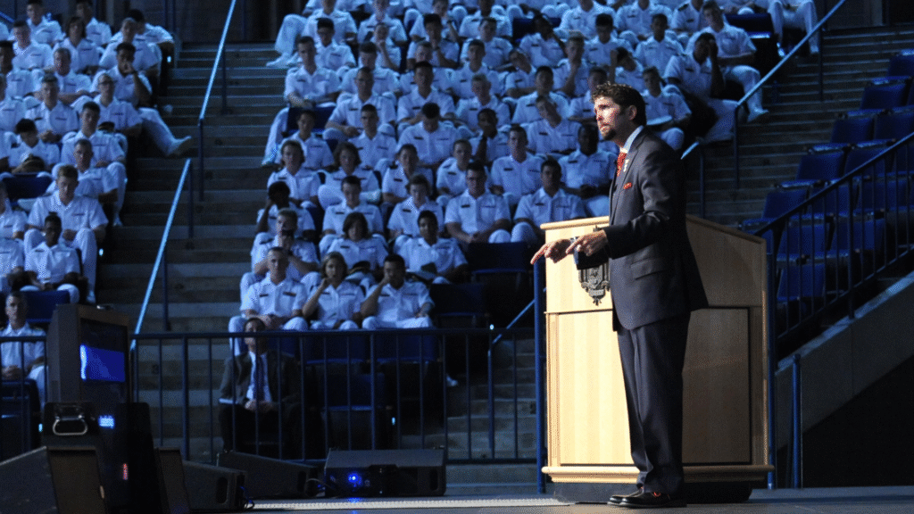 A man in a suit speaking at a podium before an audience of uniformed individuals seated in an auditorium.