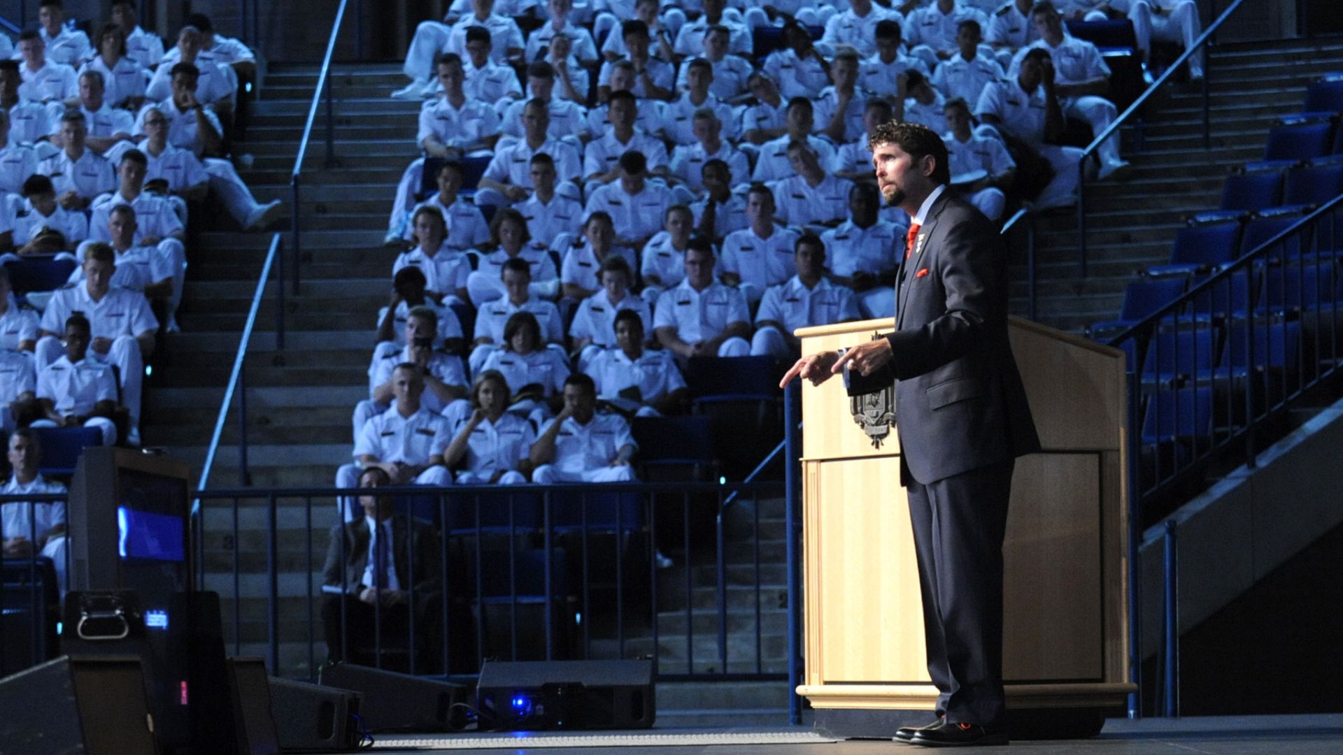 A man in a suit speaking at a podium before an audience of uniformed individuals seated in an auditorium.
