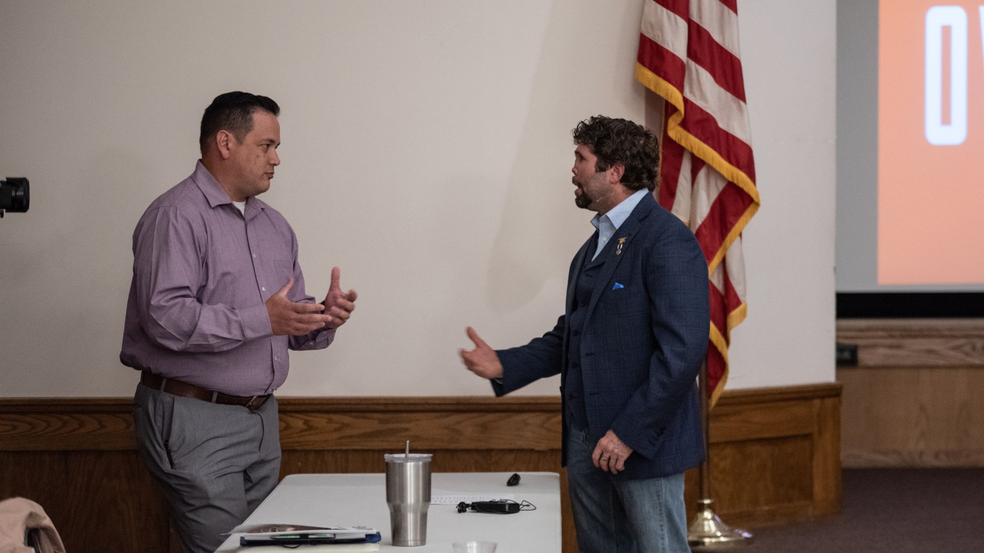 Two men gesturing during a discussion near a table with a water bottle, with an American flag and a screen in the background.