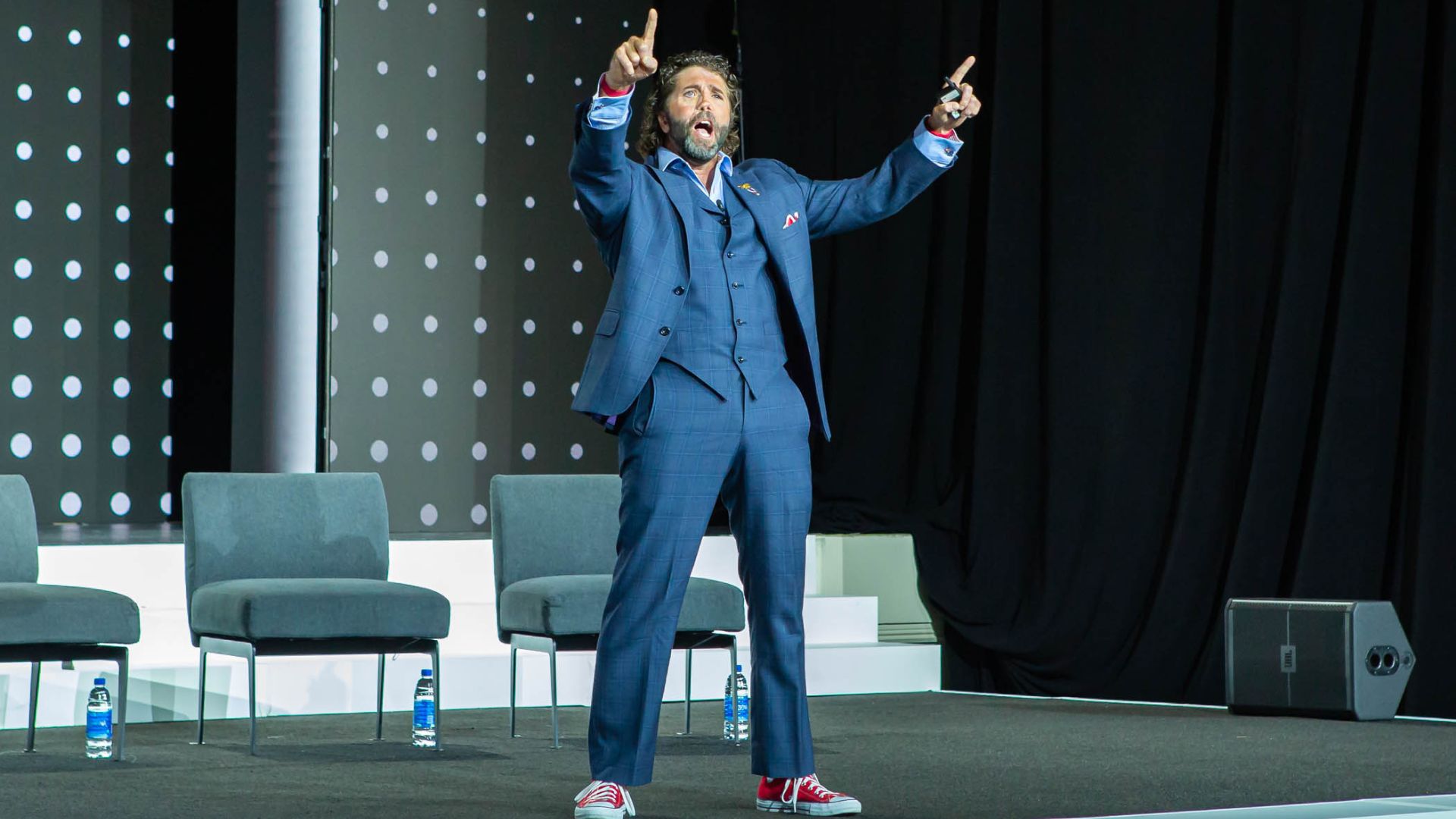 Image of a man in a blue suit and red sneakers on stage, raising both arms energetically during a keynote presentation, with chairs and water bottles in the background.