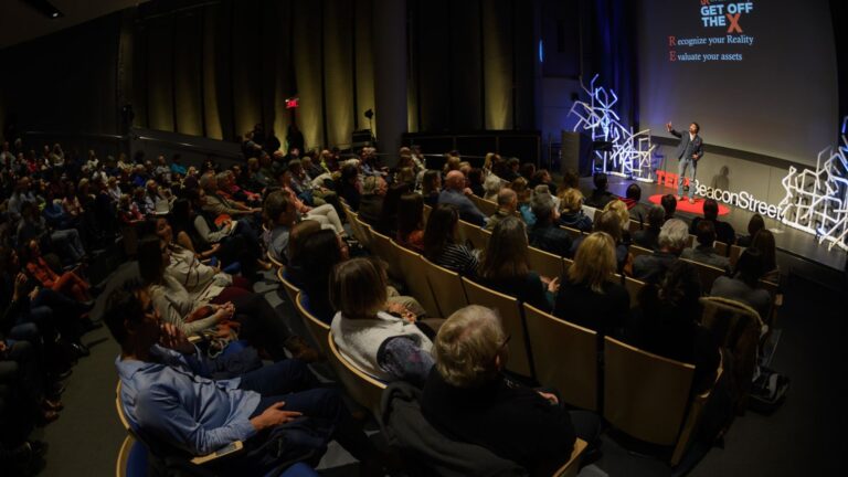 A speaker on a TEDx BeaconStreet stage addressing a large audience, with a screen displaying "Get Off" and tips to recognize and evaluate reality.