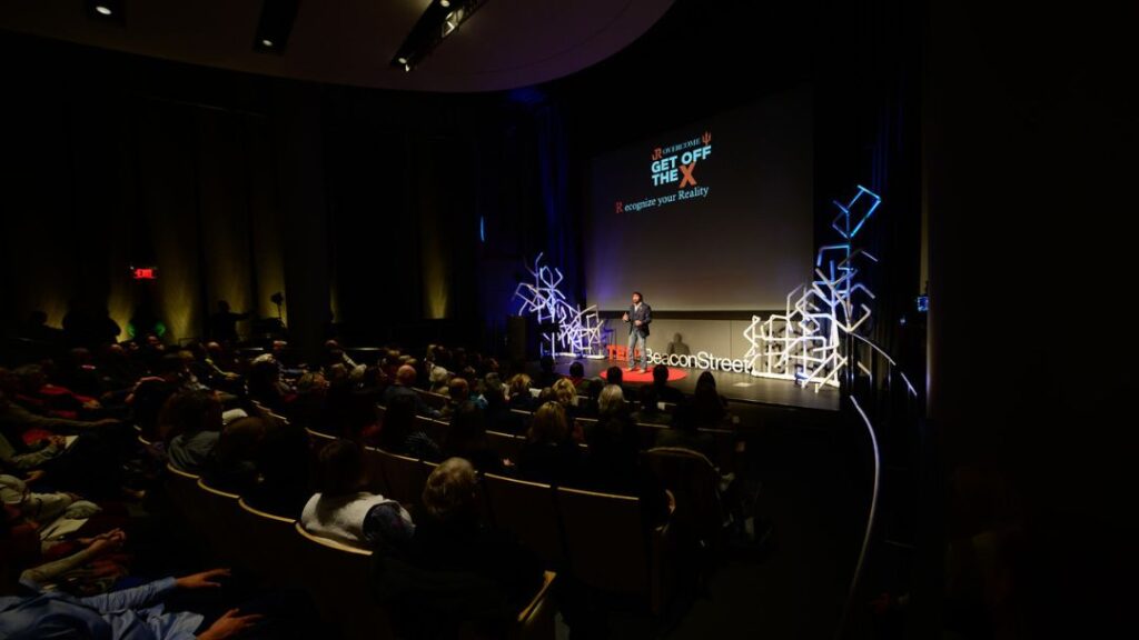 A speaker presents at an event titled "GET OFF THE X" and "Recognize your Reality" to a large audience seated in a dark auditorium.