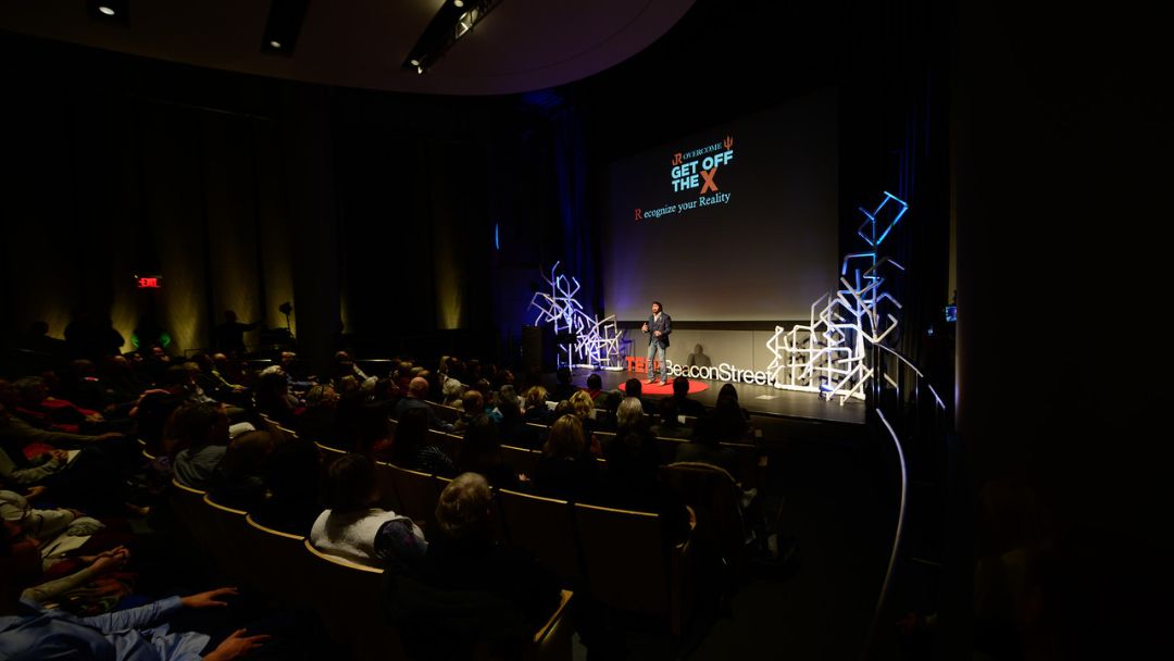 A speaker presents at an event titled "GET OFF THE X" and "Recognize your Reality" to a large audience seated in a dark auditorium.