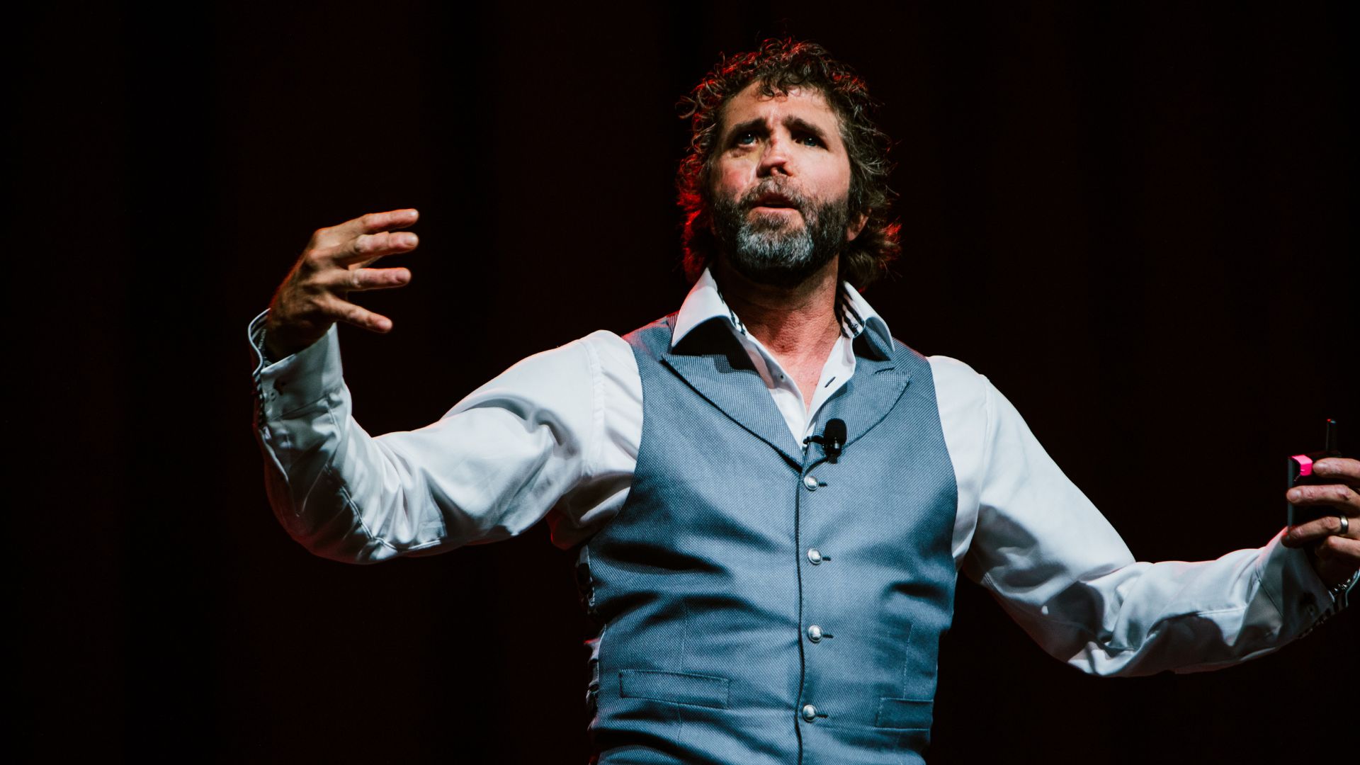 A bearded man in a white shirt and blue vest gestures dramatically on stage, holding a microphone, under warm lighting.
