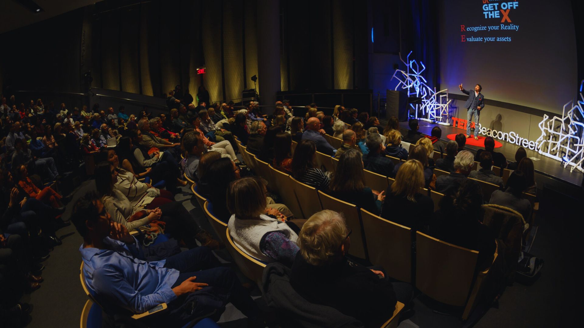 Speaker on TEDxBeaconStreet stage addresses large audience. Slide: "GET OFF THE X" – Recognize Reality, Evaluate Assets.