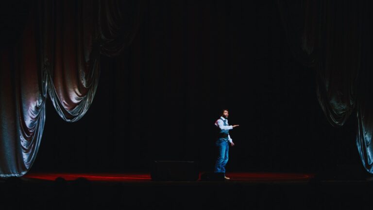 A man stands alone on a dimly lit stage, dressed in a vest, white shirt, and blue jeans, gesturing with his right hand in a wide shot.