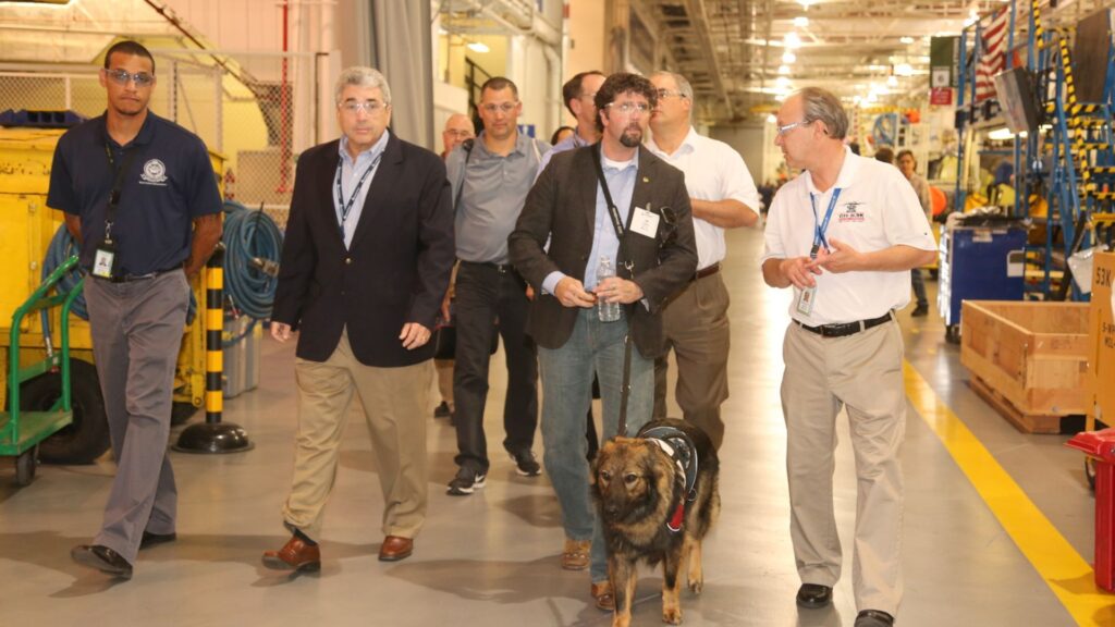 Jason Redman, retired Navy SEAL, tours a manufacturing facility with his service dog, a harnessed German Shepherd, accompanied by staff and guides in an industrial warehouse setting.