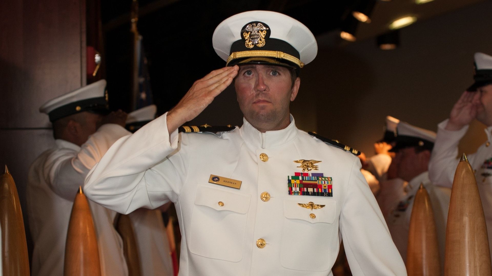 Retired Navy SEAL Lt. Jason Redman saluting in white dress uniform with SEAL Trident, ribbons, and name tag "REDMAN" during a formal ceremony.