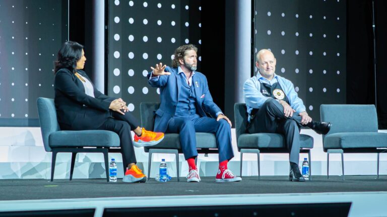 Panel of three speakers engaged in discussion on stage at conference; practical 2026 guide to getting better at public speaking.