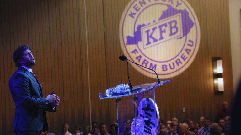 Professional speaker at podium addressing Kentucky Farm Bureau audience with organization logo projected behind him on stage.