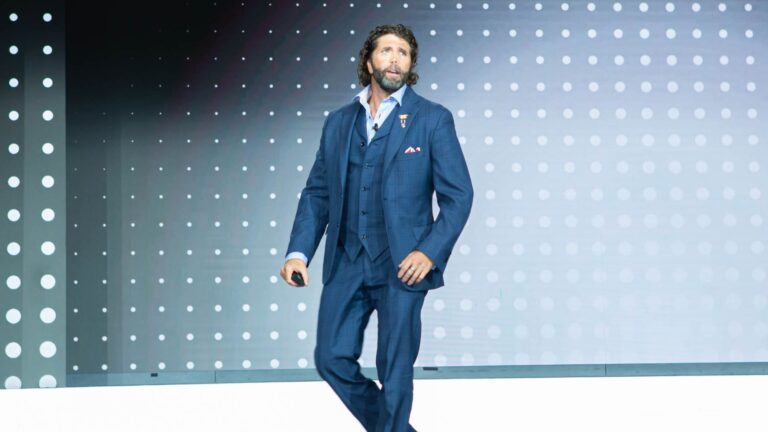 Motivational speaker in a navy suit commanding a large corporate event stage, showcasing the impact and benefits of hiring a keynote speaker for company events.