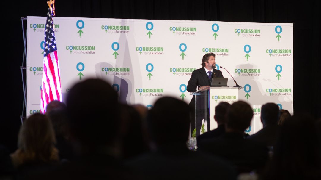 Keynote speaker presenting at a formal Concussion Legacy Foundation event before a seated audience, illustrating how different speaker types suit different event formats.